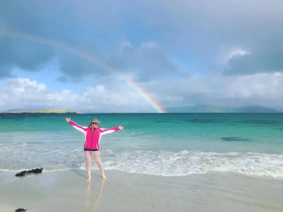 Susan on a white-sand beach with her arms open wide under a rainbow