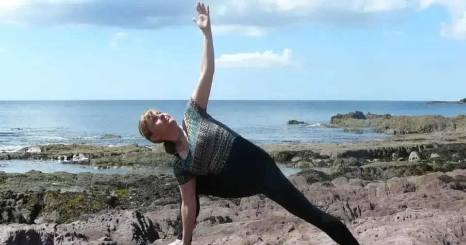 Susan in a side-angle stretch on the rocky shore, with the sea and hills behind her