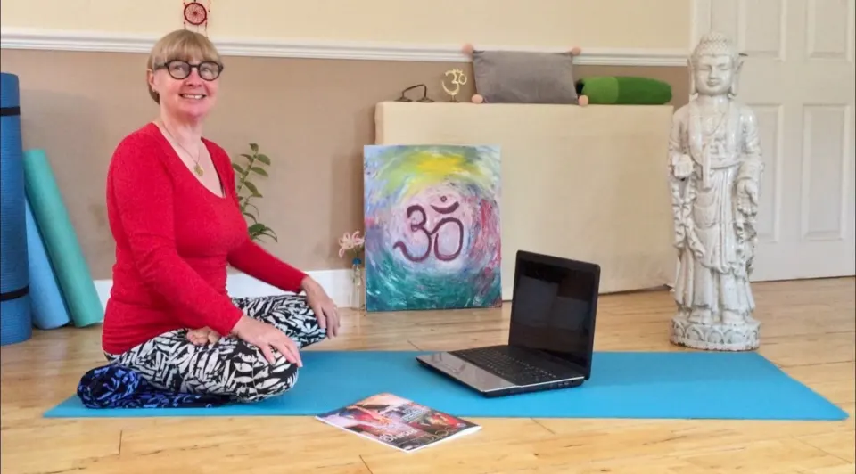 Susan sitting on her mat in her yoga room, teaching to a laptop